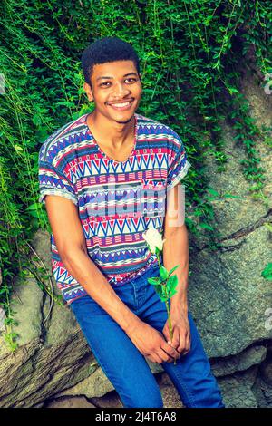 Man Waiting for You. Wearing a short sleeve, collarless, colorful pattern shirt, blue jeans, a young black guy is sitting against rocks with green lea Stock Photo