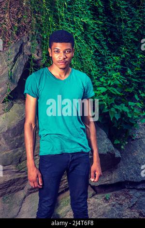 Portrait of Young Black Man. Wearing a green, short sleeve, V-neck T shirt, black pants, a young handsome guy is standing by rocks with green leaves, Stock Photo