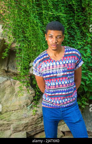 African American Man Casual Fashion in New York. Wearing colorful patterned, short sleeves, collarless shirt, blue jeans, a young guy standing by rock Stock Photo