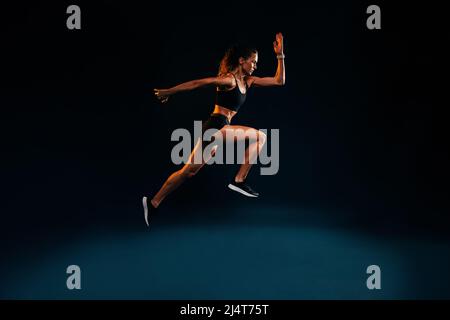 Fit woman running and jumping in the studio. Young female sprinting against dark background. Stock Photo
