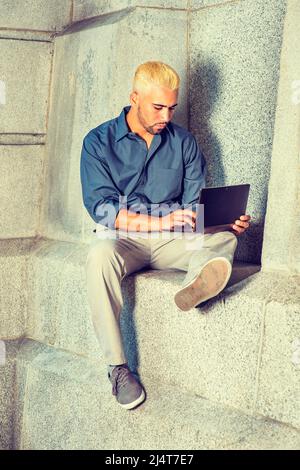 Young Man Working Outside. Wearing a blue shirt, gray pants, casual shoes, a young guy with beard, yellow hair is sitting by a concrete wall, looking Stock Photo