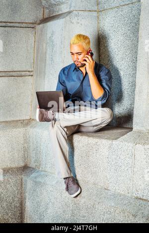 Man Working Outside. Wearing a blue shirt, gray pants, casual shoes, a young guy is sitting by a concrete wall, looking down, working on a laptop comp Stock Photo