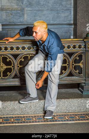 Man Looking for You. Wearing a blue shirt, gray pants, casual shoes, a young guy with beard, yellow hair is standing by old fashion style railing in a Stock Photo