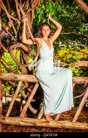 Portrait of Pretty Lady. Dressing in a light blue long dress, a young woman with long curly hair is sitting on wooden fence, raising arms, holding bra Stock Photo