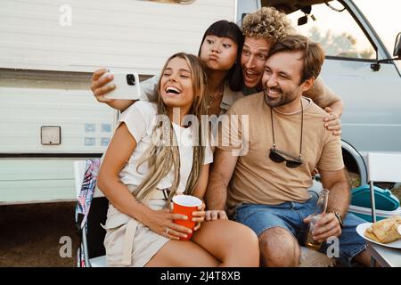 Multiracial two couples laughing and taking selfie photo while leaning on trailer outdoors Stock Photo