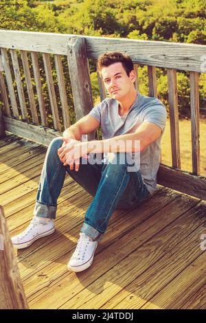 Man Relaxing Outside. Wearing a gray T shirt, jeans, white sneakers, a young handsome guy is sitting on the wooden floor, back against fence in a remo Stock Photo