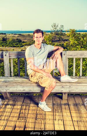 Man Relaxing Outside. Wearing a gray T shirt, yellow pants, white sneakers, a young handsome guy is sitting on wooden bench against fence in remote lo Stock Photo
