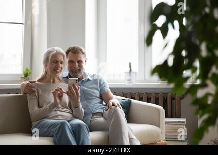 Smiling mature couple using cellphone seated on sofa at home Stock Photo