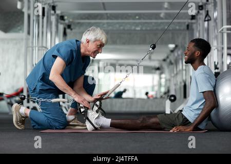 Portrait of smiling senior therapist helping young man with rehabilitation exercises in gym, copy space Stock Photo