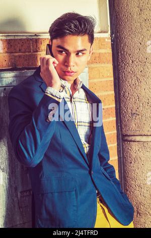 Man Calling Outside. Dressing in blue blazer, yellow pants, hand holding a mobile phone. a young handsome guy is standing by a brown brick wall, looki Stock Photo