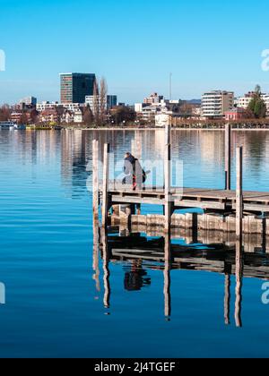 Zug, Switzerland - December 31, 2021: Young woman on a wooden pier is taking photo of the cityscape of Zug, a political municipality and capital of th Stock Photo