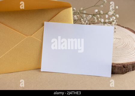 Rustic invitation or greeting card mock up template with golden envelope, blank empty white paper, wood and flowers on craft paper. Stock Photo