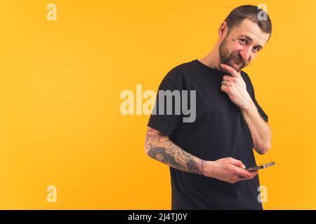 Smiley friendly bearded caucasian guy with tattoos holding smartphone in one hand and touching his beard with the other while looking at camera. Isolated studio shot over yellow background. High quality photo Stock Photo
