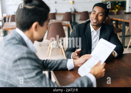 Two positive multiethnic coworkers sitting together at desk in modern office room and discussion over documents. Stock Photo