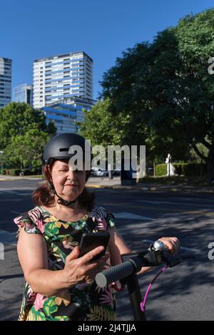 Latin mature woman using a protective helmet looking at her smartphone before riding her electric kick scooter. Concepts of tech, active retired life, Stock Photo