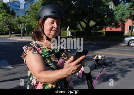 Latin mature woman using a sports helmet looking at her smart phone before riding her electric kick scooter. Concepts of technology, healthy and activ Stock Photo