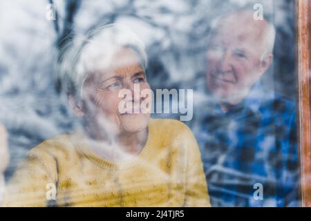 Shot from behind the window glass. Concerned gray-haired elderly woman looking out of the window, while her husband in blue flannel shirt is standing behind her. High quality photo Stock Photo