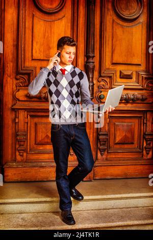 Busy Student. Wearing a patterned sweater, red tie, blue jeans, leather shoes, a young guy is walking down from office, talking on his mobile phone, w Stock Photo