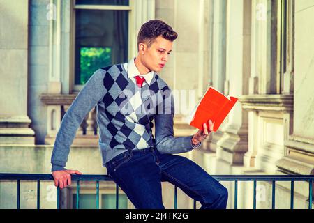 Man Reading Outside.  Dressing in a black, white, gray patterned sweater, jeans,  a young handsome college student is sitting on a railing in office b Stock Photo