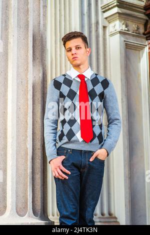 Portrait of College Student. Dressing in a black, white, gray patterned sweater, red tie, jeans, a young handsome businessman is standing by a pillar Stock Photo