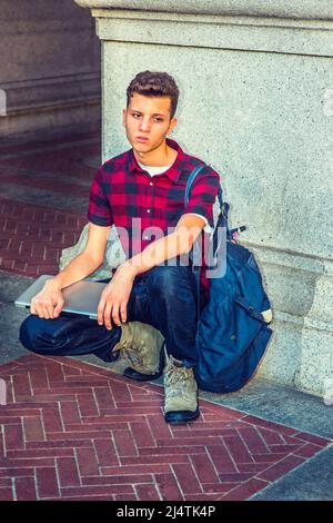 Dressing in a short sleeve, black, red patterned shirt, jeans, boot shoes, carrying shoulder bag, holding a laptop computer, a young guy is squatting Stock Photo