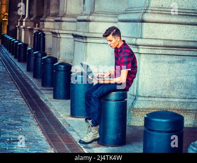 Man Working on street. Wearing a short sleeve, patterned shirt, jeans, boot shoes, holding a laptop computer, a young guy is sitting on a metal pillar Stock Photo