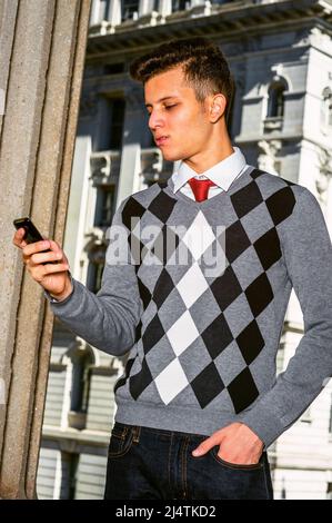 Technology in our daily life. Wearing black, white, gray patterned sweater, red necktie, jeans, a young college student checking messages on his mobil Stock Photo
