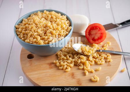 Raw or uncooked Elbow Pasta in a blue bowl and on a wooden chopping board with a spoon on the side Stock Photo