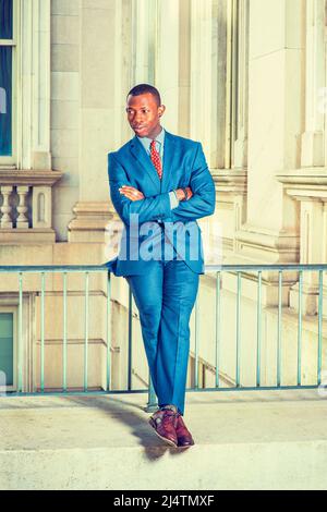 Dressing formally in blue suit, patterned undershirt, tie, leather shoes, short haircut, crossing arms and legs, a young black businessman is sitting Stock Photo