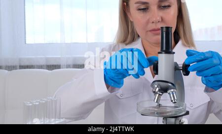 Female laboratory assistant analyzes samples using a microscope while working at a table in a laboratory, doing research Stock Photo