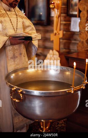 Unrecognizable bearded priest standing at font decorated with candles, reading prayer book during ceremony of baptism. Stock Photo