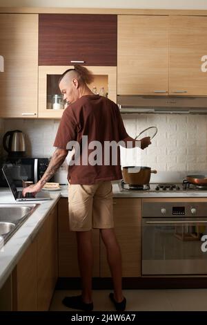 Young man in loungewear cooking soup for dinner and checking e-mails and notifications on laptop Stock Photo