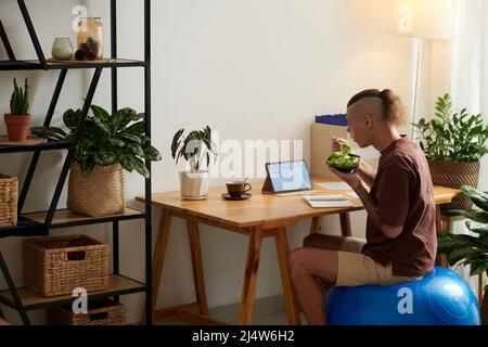 Project manager sitting on fitness ball, eating salad and checking mock-ups sent by UI designr Stock Photo