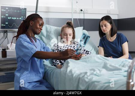 Healthcare clinic pediatric smiling nurse showing unwell little girl radiography results. Mother and surprised daughter looking at x-ray scan image while in patient recovery ward room. Stock Photo