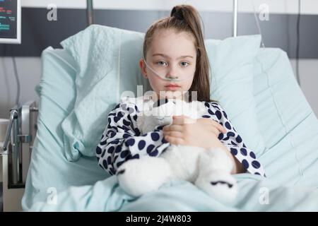 Sick kid holding teddybear and wearing oxygen tube while resting alone in pediatric clinic bed. Unhealthy child in healthcare pediatric hospital patients treatment ward room while looking at camera. Stock Photo