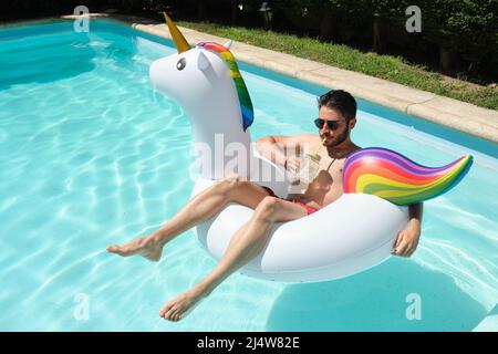Young man with sunglasses drinks a refreshment on a unicorn inflatable ring. Stock Photo