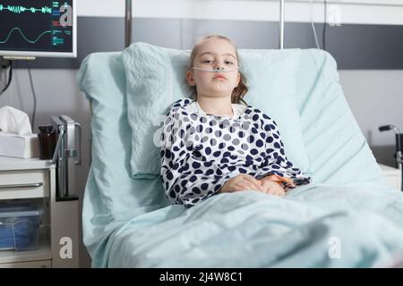 Lonely young pale sick girl patient resting in pediatric clinic bed while in recovery ward room. Ill child sitting alone in hospital bed while wearing nasal oxygen tube for life support. Stock Photo