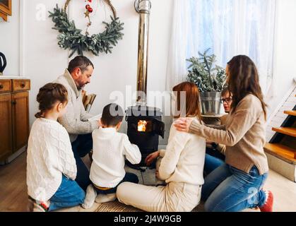 Big family dressed in cozy sweaters sitting near the fireplace in the house in the mountains durnig winter time together. Holiday and travel concept Stock Photo