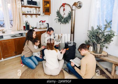 Big family dressed in cozy sweaters sitting near the fireplace in the house in the mountains durnig winter time together. Holiday and travel concept Stock Photo