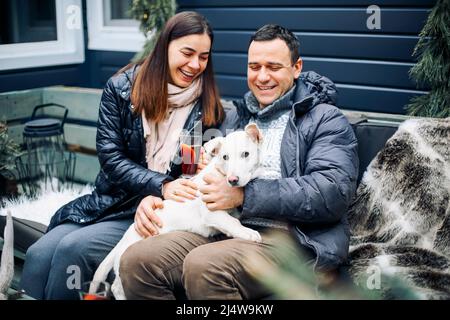 Young couple hugs their white cute pet dog. Man and woman at new year fair, drinking mulled red wine, happy and smiling. Stock Photo