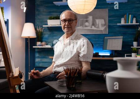 Portrait of inspired elderly man posing confident holding pencil in professional art studio sitting in front of easel. Senior sketch artist feeling creative at table with vase model and hb pencils. Stock Photo