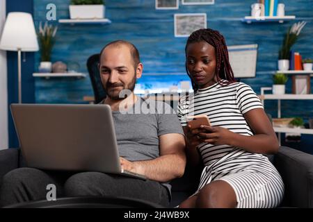 Happy caucasian man websurfing on laptop while african american woman holding smartphone and looking at computer screen. Couple sitting on sofa in living room checking bills and financial issues on computer and phone Stock Photo