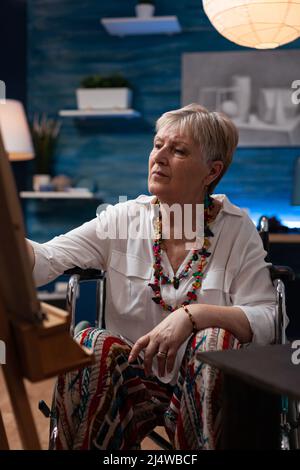 Portrait of older woman drawing sketch using pencil on canvas in front of easel in art studio. Retired art teacher using wheelchair doing creative freehand black and white drawings. Stock Photo