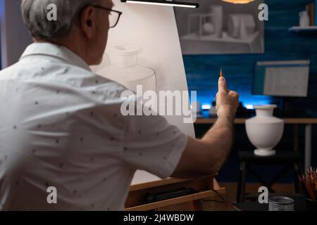 Focus on hand of creative senior artist looking at plaster vase measuring proportions using pencil doing creative pencil drawing. Elderly man in the evening sketching scale model in home art studio. Stock Photo