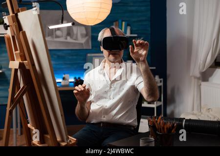 Retired art teacher with modern technology vr goggles looking at 3d artwork in creative workshop. Senior sketch artist using virtual reality glasses doing air gesture improving freehand technique. Stock Photo