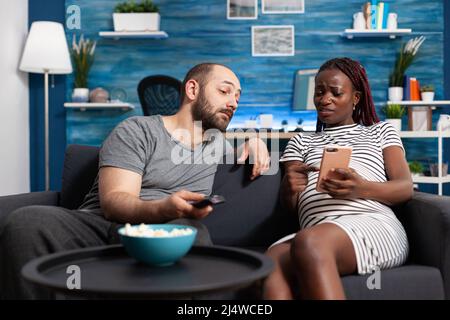 Worried african american pregnant woman sitting on sofa at home while showing curious caucasian man something on smartphone. Multiethnic young couple looking at wrong issued bills using modern phone. Stock Photo