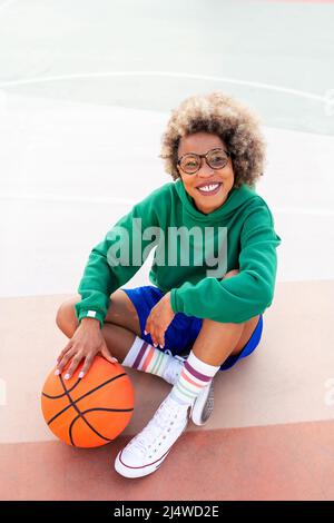 smiling latin woman looking at camera sitting on the court after basketball practice, concept of urban sport outdoors, copy space for text Stock Photo