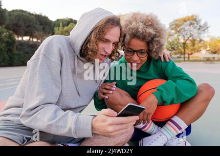 man and woman have a good time looking at things on the cell phone while sitting after basketball practice in a city park, concept of friendship and u Stock Photo