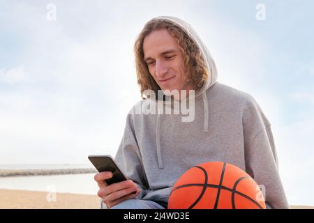 smiling caucasian man in a hoodie using his phone next to the beach with a basketball in his hand, concept of lifestyle and technology Stock Photo
