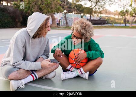 man and woman rest and have fun looking at the phone after basketball practice in a city park, concept of friendship and urban sport in the street, co Stock Photo